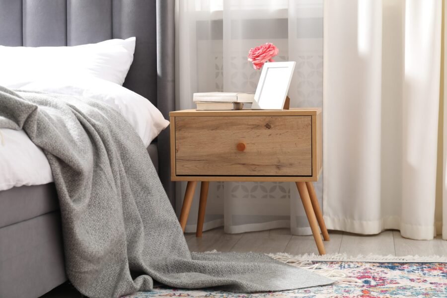 Bedside table with books, flower and frame near bed indoors